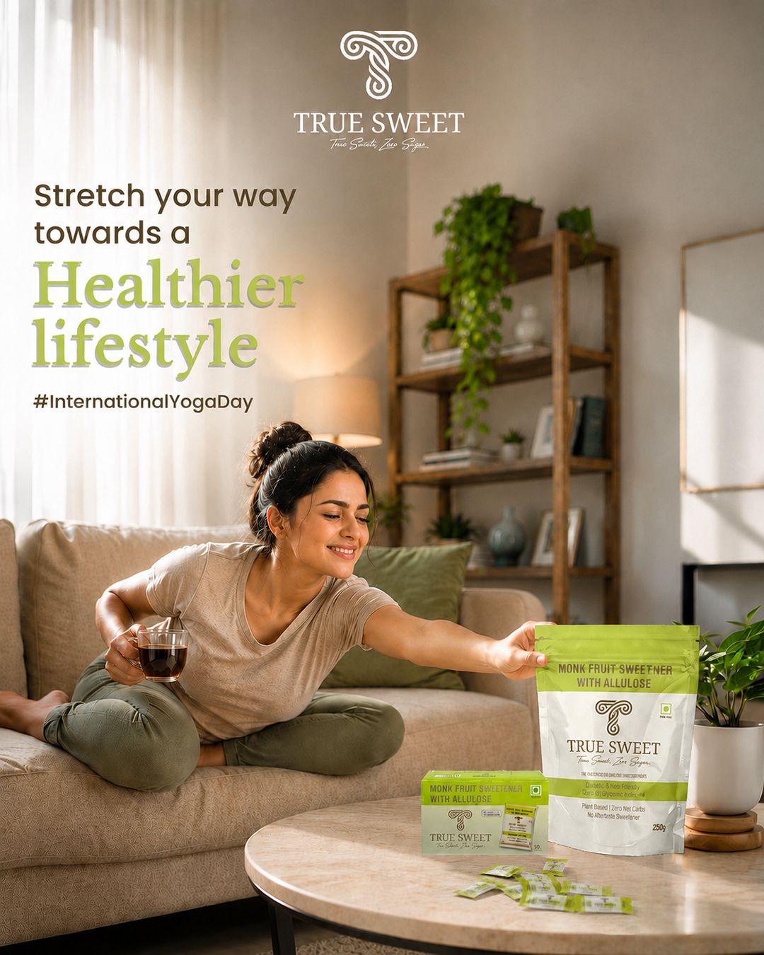A woman sits on a beige sofa in a bright living room, holding a glass of dark tea while reaching toward a bag of TRUE SWEET Non-GMO Fruit Sweeteners. The bag is white and light green, and the scene includes promotional text for a healthier lifestyle and International Yoga Day.